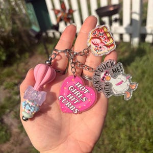 May include: A keychain featuring a pink hot air balloon bead, a translucent purple bow bead, a blue and white patterned bead, a pink heart charm with the text HOT GIRLS HIT CURBS, a glittery elephant charm, and a duck charm with the text DON'T DUCK WITH ME.
