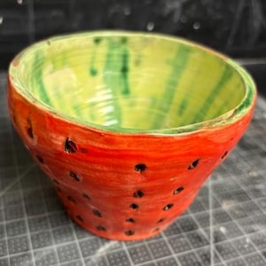 May include: A handmade ceramic bowl, painted to resemble a watermelon. The exterior is red with black seed-like markings, while the interior is green and yellow. The bowl is a unique piece of kitchenware.