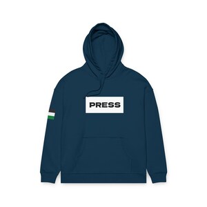 Może przedstawiać: Granatowa bluza z kapturem z napisem "PRESS" w białym prostokącie na piersi. Mały wzór flagi znajduje się na lewym rękawie. Bluza ma kaptur i przednią kieszeń.
