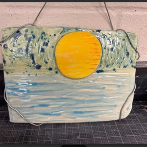 May include: Handmade ceramic wall art depicting a sunset over the ocean. The artwork features a large yellow sun, blue water, and a textured sky with blue accents. The piece is rectangular and has a wire for hanging.