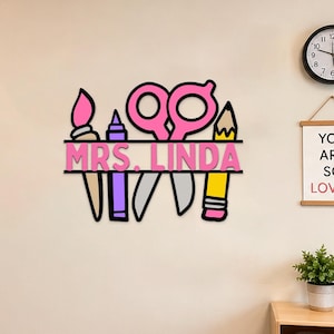 Puede incluir: Un letrero con el nombre de un profesor en rosa y negro con el texto "MRS. LINDA" y útiles escolares. El letrero incluye un pincel, tijeras, un lápiz y otros útiles escolares. El letrero está montado en una pared beige.