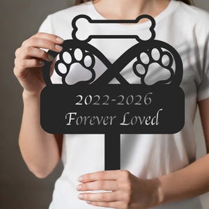 May include: Black metal pet memorial stake featuring paw prints, a bone, and the text "2022-2026 Forever Loved." The stake is designed to be placed in the ground as a tribute to a beloved pet.