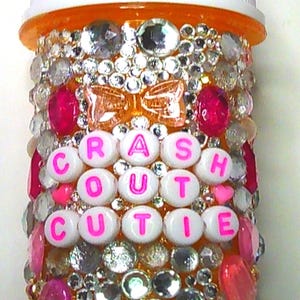 May include: A bedazzled pill bottle with the word "RASH CUTIE" spelled out in pink letters. The bottle is covered in rhinestones, pink and clear gems, and a pink bow.