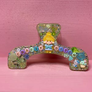 May include: A resin hair claw clip decorated with glitter, small beads, and charms. The center features a yellow-haired character figurine. The text CRAFTY BITCH is spelled out in colorful letter beads across the front, accented by miniature food and flower charms.