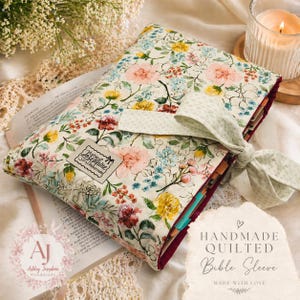 May include: A handmade, quilted Bible sleeve featuring a floral design in pink, yellow, and blue. It has a burgundy border and a light green bow closure. The text "Ashley Josephine Handmade" is on a small tag.