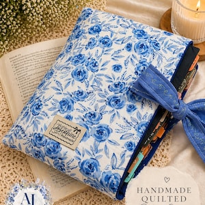 May include: A handmade quilted Bible sleeve with a blue floral pattern on a white background. The sleeve has a blue bow and a label that says "Ashley Josephine Handmade". The text "HANDMADE QUILTED Bible Sleeve" is also visible.