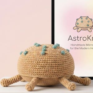 May include: A tan crochet microbe plush toy with small teal accents on its rounded top and several short legs. Behind it, a tablet displays the text AstroKnit Handmade Microbes for the Modern Home with a matching illustration of the microbe.