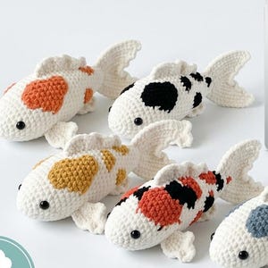 Könnte beinhalten: Fünf handgefertigte Häkel-Koikarpfen mit orangefarbenen, schwarzen, gelben und blauen Flecken auf weißen Körpern. Ein Tablet zeigt den Text CROCHET KOI FISH PATTERN: A Complete Beginner's Guide sowie ein Symbol für digitalen Download.