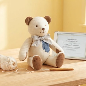 Puede incluir: Un oso de peluche de pana color crema con orejas y almohadillas marrones, que lleva una pajarita a rayas y un pequeño bolsillo de mezclilla azul. Junto a él hay un ovillo de lana crema, un ganchillo de madera y una tableta con un certificado de creación.