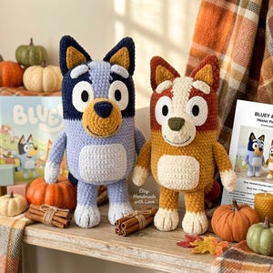 May include: Two handmade crochet plush toys, Bluey and Bingo, stand side-by-side. Bluey is blue and Bingo is orange. The scene includes pumpkins, candles, a book, and a pattern sheet. The toys are handmade with love.