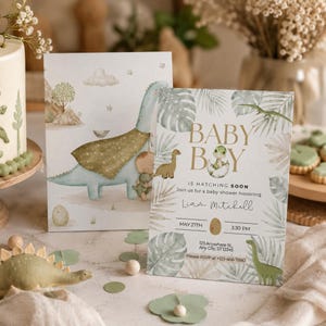 Puede incluir: Una tarjeta de invitación para baby shower con una ilustración en acuarela de un dinosaurio verde saliendo de un huevo, rodeado de hojas tropicales. El texto dice BABY BOY IS HATCHING SOON, para Liam Mitchell, 27 de mayo, 3:30 PM.