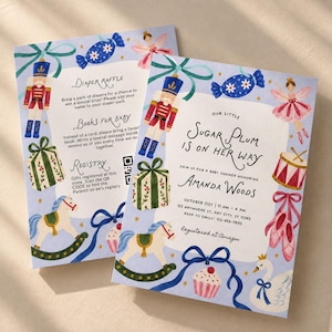 Puede incluir: Dos tarjetas de invitación rectangulares para baby shower con ilustraciones en acuarela de cascanueces, caballitos de madera, zapatillas de ballet y regalos sobre un fondo azul claro. El texto incluye detalles para Amanda Woods y el registro.