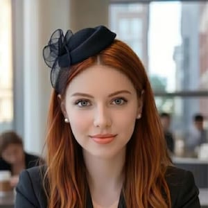 Puede incluir: Un sombrero negro con un detalle transparente y con volantes se lleva en la cabeza. El sombrero está colocado sobre un pelo largo y castaño rojizo. La persona lleva una blazer negra. El fondo está borroso, lo que sugiere un entorno interior.