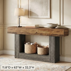 May include: A rustic console table with a distressed wood top and gray supports. The table has two woven baskets and a lower shelf. Dimensions: 11.8"D x 63"W x 32.3"H. A lamp, books, and a bowl are on the table.