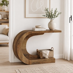 May include: A C-shaped wooden console table with a natural wood grain finish. The table features a flat top surface holding a beige ceramic vase with green foliage, a small bowl, and two books. A woven basket with a cloth sits on the base.