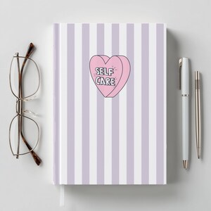 Op de afbeelding: Een notitieboek met een gestreepte lavendel en witte kaft, met een roze hartvormige afbeelding met de tekst "SELF CARE". Het notitieboek wordt vergezeld door een bril en twee pennen.