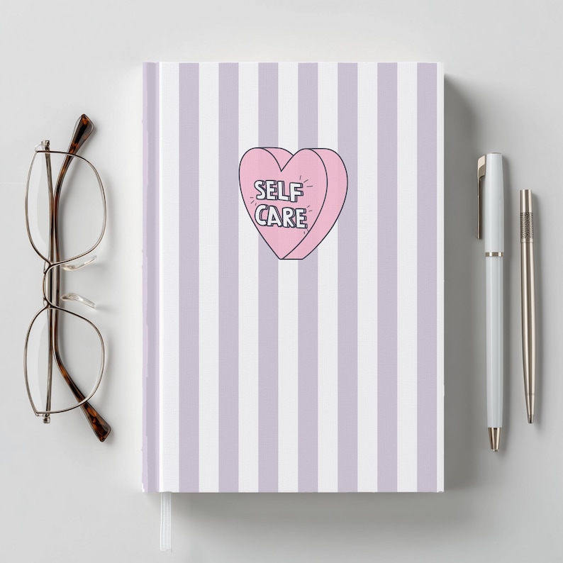 Op de afbeelding: Een notitieboek met een gestreepte lavendel en witte kaft, met een roze hartvormige afbeelding met de tekst "SELF CARE". Het notitieboek wordt vergezeld door een bril en twee pennen.