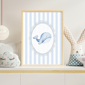 Op de afbeelding: Een ingelijste print met een aquarel illustratie van een lachende blauwe walvis in een ovale lijst. De achtergrond heeft verticale blauwe en witte strepen. Het kunstwerk wordt tentoongesteld op een witte plank met decoratieve items.