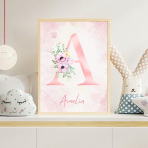 May include: A framed pink watercolor print featuring the letter "A" adorned with floral accents. The name "Amelia" is written in cursive below the letter. The background has a soft, pink watercolor wash.