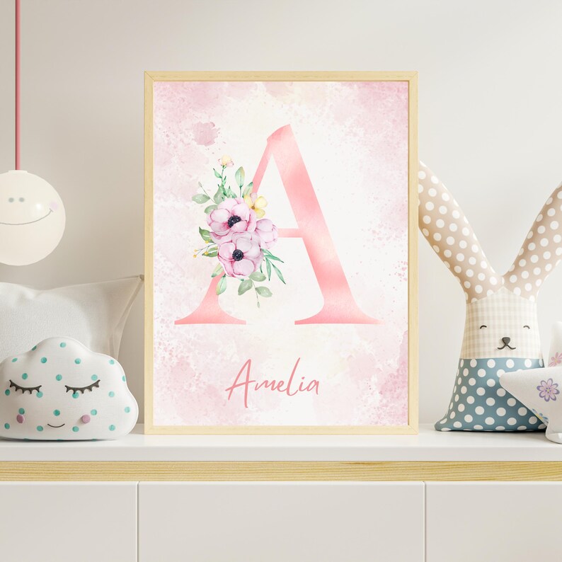 May include: A framed pink watercolor print featuring the letter "A" adorned with floral accents. The name "Amelia" is written in cursive below the letter. The background has a soft, pink watercolor wash.