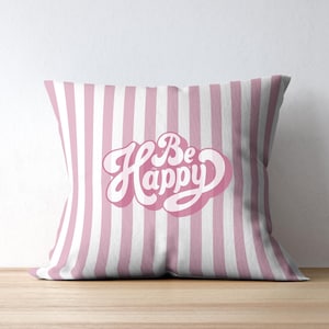 Op de afbeelding: Een vierkant decoratief kussen met verticale roze en witte strepen. Het kussen toont de woorden "Be Happy" in een cursief lettertype met een roze omtrek.