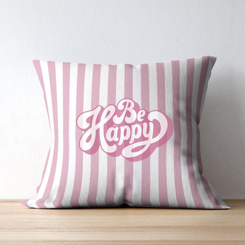 Op de afbeelding: Een vierkant decoratief kussen met verticale roze en witte strepen. Het kussen toont de woorden "Be Happy" in een cursief lettertype met een roze omtrek.