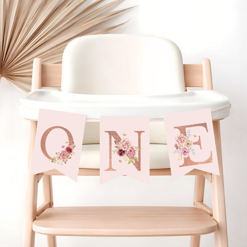 May include: A banner spelling "ONE" in rose gold glitter letters, each adorned with floral arrangements. The banner is displayed on a light wood high chair, with a neutral background. The letters are on pink pennants.