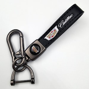 May include: Black leather keychain with a metal clasp and a Cadillac logo. The keychain features the Cadillac logo in the center and the word "Cadillac" embroidered in white. The metal clasp is gunmetal gray.