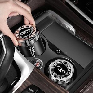 Könnte beinhalten: Zwei zylindrische, schwarze Auto-Lufterfrischer mit metallisch silbernen turbinenartigen Oberteilen, die das Audi-Logo tragen. Die Seite einer Einheit zeigt den Text CAR PERFUME AIR PAMTI CAT. Diese Geräte sind für den Getränkehalter konzipiert.