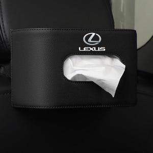 Puede incluir: Un soporte rectangular negro para pañuelos de coche hecho de material texturizado con el logotipo y el texto de Lexus en plata impresos en la parte frontal. Un pañuelo blanco sobresale por la abertura ovalada central del soporte, montado en un asiento.