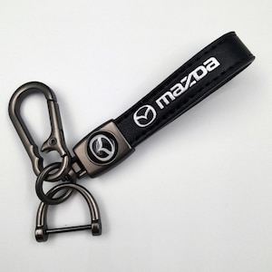 Puede incluir: Llavero de cuero negro con el logotipo y texto Mazda en blanco. El llavero tiene un cierre y herrajes de color metalizado. El logotipo de Mazda también aparece en los herrajes metálicos.