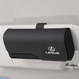 Peut inclure: Un étui à lunettes rectangulaire noir pour pare-soleil de voiture, arborant le logo Lexus blanc sur le rabat avant. L'étui est fixé à un pare-soleil de véhicule gris clair par une sangle noire assortie, offrant un rangement pour les lunettes.
