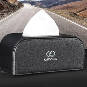 Peut inclure: Un porte-boîte à mouchoirs rectangulaire noir avec une surface texturée et des bords arrondis. L'avant présente un logo Lexus argenté et le mot LEXUS imprimé en blanc. Un mouchoir blanc est tiré vers le haut depuis l'ouverture centrale.