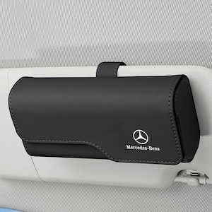 Puede incluir: Un estuche rectangular negro para gafas con cierre de solapa magnética, que presenta el logotipo y el texto de Mercedes-Benz en blanco en la parte frontal. El estuche está sujeto a un parasol de coche gris claro mediante una pequeña correa.