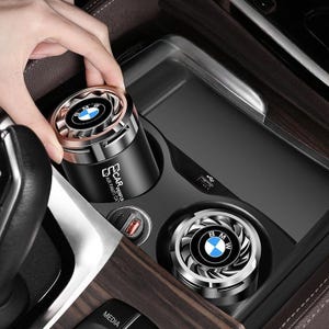 May include: Two cylindrical black car air fresheners with metallic silver fan-style tops featuring the BMW logo. The units are designed to fit into a vehicle cup holder, with one unit placed in the holder and the other being positioned by hand.