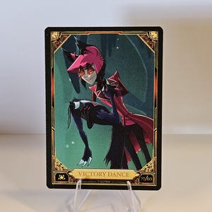 May include: A trading card featuring a character in a red and black outfit, with a red and black color scheme. The card has the text "VICTORY DANCE" and the number "77/60". The card is displayed on a clear plastic stand.