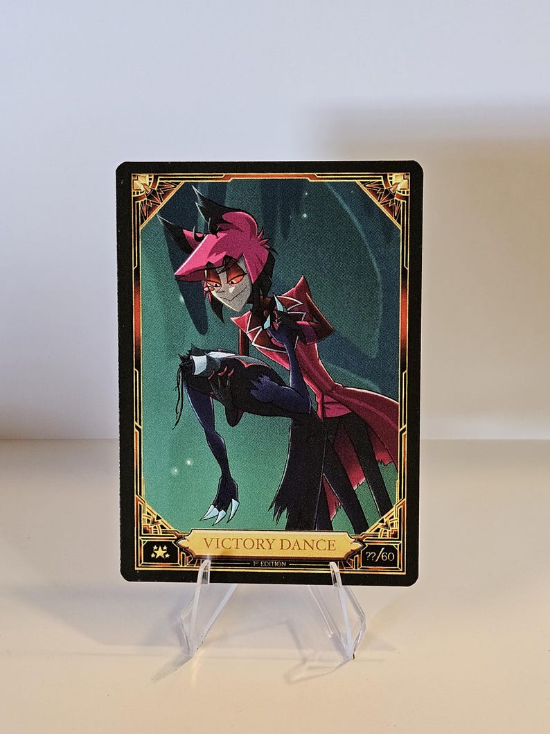 May include: A trading card featuring a character in a red and black outfit, with a red and black color scheme. The card has the text "VICTORY DANCE" and the number "77/60". The card is displayed on a clear plastic stand.