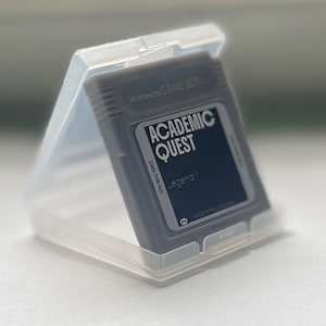 Puede incluir: Un cartucho gris de Nintendo Game Boy con la etiqueta "ACADEMIC QUEST" en una funda de plástico transparente. El cartucho tiene una etiqueta negra con texto blanco y un fondo azul oscuro. La funda está abierta, mostrando el cartucho en ángulo.