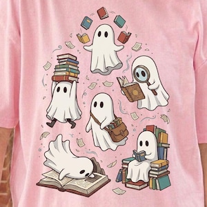 May include: A light pink t-shirt featuring a graphic print of five white ghosts interacting with books. One ghost balances a stack of books, another uses a magnifying glass, one carries a satchel, one sits on a pile of books, and one lies on an open book.