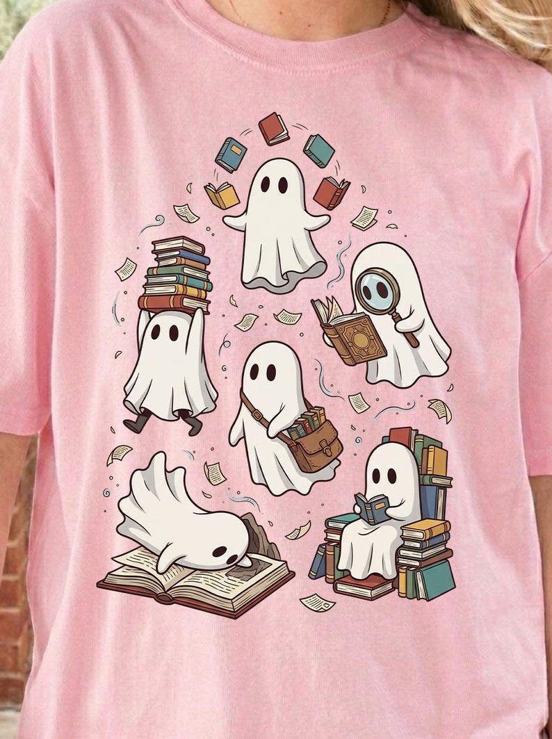 May include: A light pink t-shirt featuring a graphic print of five white ghosts interacting with books. One ghost balances a stack of books, another uses a magnifying glass, one carries a satchel, one sits on a pile of books, and one lies on an open book.