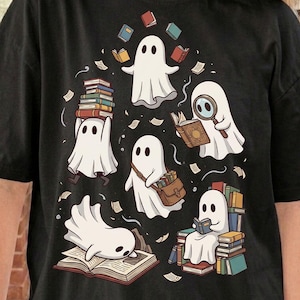 May include: A black t-shirt featuring a graphic illustration of five white ghosts interacting with books. One ghost carries a stack of books, another uses a magnifying glass, one wears a satchel, one reads, and one lies on an open book.