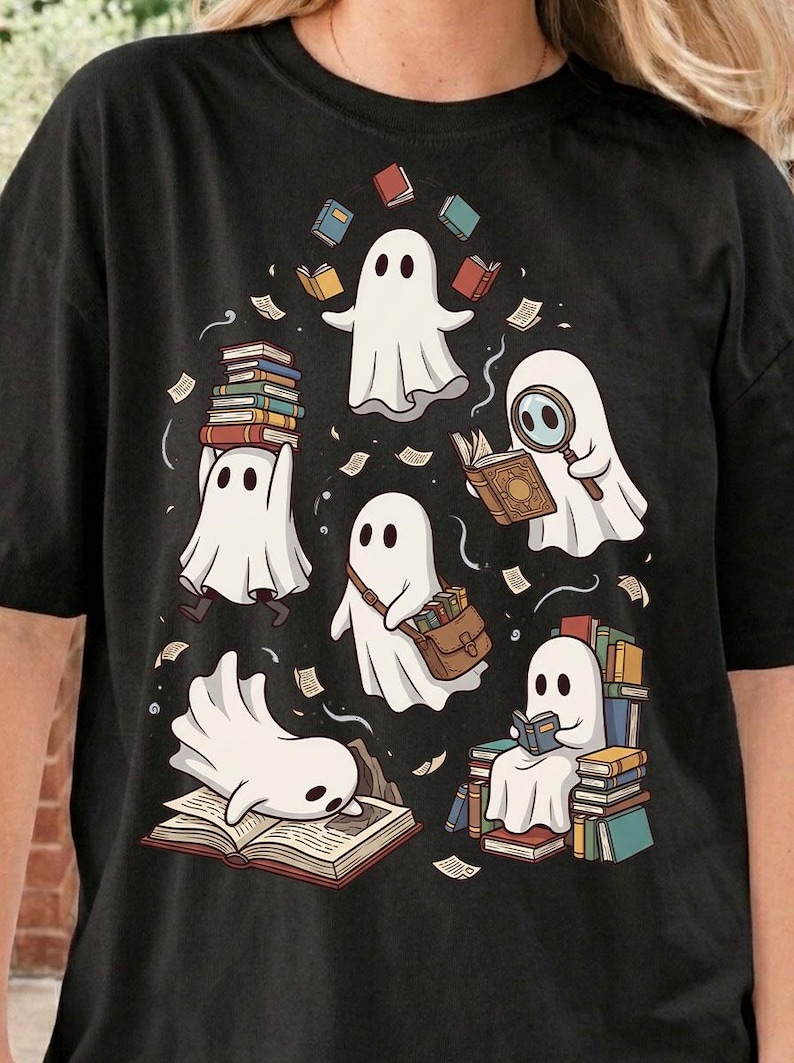 May include: A black t-shirt featuring a graphic illustration of five white ghosts interacting with books. One ghost carries a stack of books, another uses a magnifying glass, one wears a satchel, one reads, and one lies on an open book.