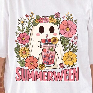 May include: A white t-shirt featuring a graphic of a ghost wearing a flower crown and holding a glass of pink fruit-infused drink. The design includes colorful flowers, leaves, and hummingbirds, with the text "SUMMERWEEN" printed in pink below.