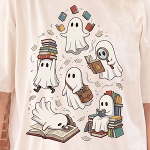 May include: A cream-colored t-shirt featuring a graphic print of five white ghosts interacting with books. One ghost balances a stack of books, another uses a magnifying glass, one carries a satchel, one sits on a pile of books, and one lies on an open book.