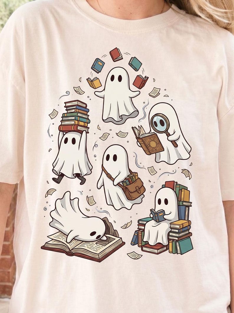 May include: A cream-colored t-shirt featuring a graphic print of five white ghosts interacting with books. One ghost balances a stack of books, another uses a magnifying glass, one carries a satchel, one sits on a pile of books, and one lies on an open book.