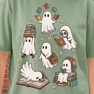 May include: A sage green t-shirt featuring a graphic illustration of five white ghosts interacting with books. One ghost juggles books, another reads with a magnifying glass, one carries a satchel, one sits on a stack of books, and one lies on an open book.