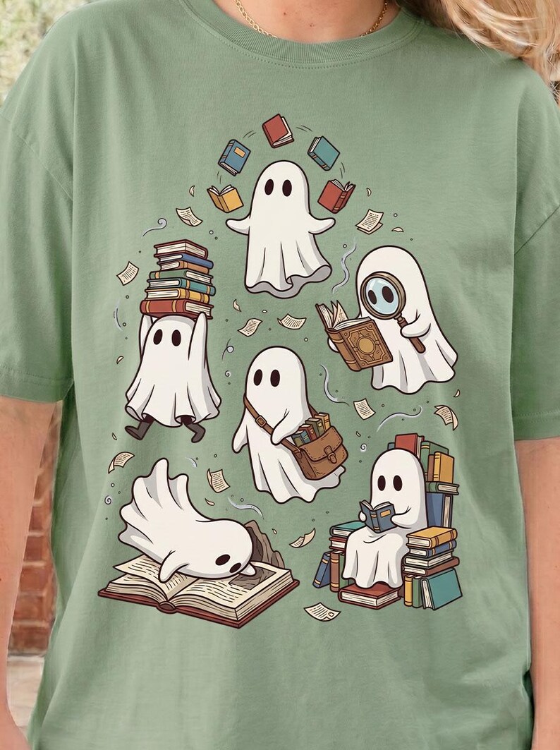 May include: A sage green t-shirt featuring a graphic illustration of five white ghosts interacting with books. One ghost juggles books, another reads with a magnifying glass, one carries a satchel, one sits on a stack of books, and one lies on an open book.