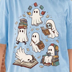 May include: A light blue t-shirt featuring a graphic illustration of five white ghosts interacting with books. One ghost balances a stack of books, another uses a magnifying glass, one carries a satchel, one reads while sitting on a pile, and one lies on an open book.