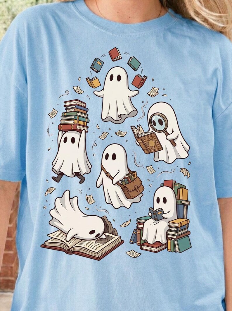 May include: A light blue t-shirt featuring a graphic illustration of five white ghosts interacting with books. One ghost balances a stack of books, another uses a magnifying glass, one carries a satchel, one reads while sitting on a pile, and one lies on an open book.