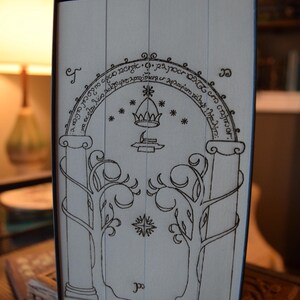 May include: A black and white illustration of a door with an archway, crown, and trees. The design is reminiscent of a fantasy theme. The image is on a light-colored surface, possibly a book cover or box.